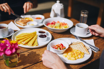 Breakfast for couple in tourist resort. Scrambled eggs and toast bread, pancakes, fruit and drinks on wooden table..