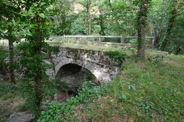 Fototapeta na wymiar Pont romain surplombant une petite rivière ombragée par de nombreux arbres sur le chemin de randonnée du ravin de Quinsat dans le puy de dôme