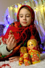 A beautiful girl is sitting in a beautiful painted scarf with a traditional pattern, the child is playing with a matryoshka. Slavic outfit and wooden toy.