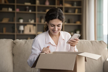 Cheerful client girl receiving purchase from internet store, using app on mobile phone, giving...