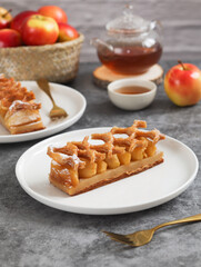 A white plate of traditional caramelize French apple tart, topped with crusty puff pastry pie. With its fruit in wooden basket and fork. Pot and cup of tea in background. Made with typical puff pastry