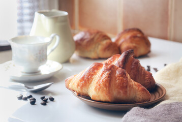 A cooked French croissant in a  plate with a cup of coffee in background. Sun light from window in the morning. It is a buttery, flaky pastry. Made with a yeast leavened dough 