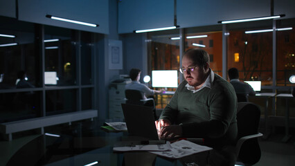 Portrait of fat businessman type on laptop in modern office at night