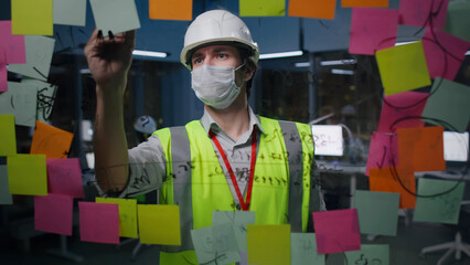 Engineer in hardhat and safety mask write on glass board with sticky notes in office