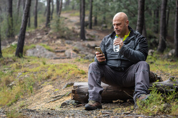 Hiker man resting on a log with a bottle of water and a mobile phone in his hands.