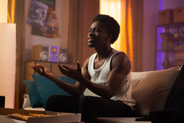 Fototapeta premium Afro man sits on the couch in the afternoon wearing a strapless T-shirt, guy eats pizza for lunch, watches TV, stretches arms out in front of him, gestures not understanding the scene
