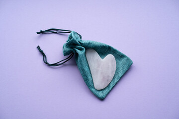 a rose quartz gouache facial massage scraper lies on a green cotton gift bag on a purple background...