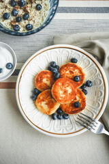 Healthy breakfast in a rustic style, oatmeal porridge, Cheesecakes or pancakes with sour cream and fresh blueberries on a linen tablecloth.
