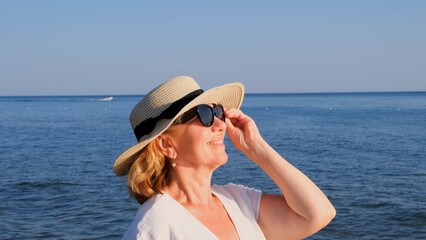 beautiful 50 year old woman in a straw hat and sunglasses on a blue sea background. Summer, vacation, vacation, active retirees