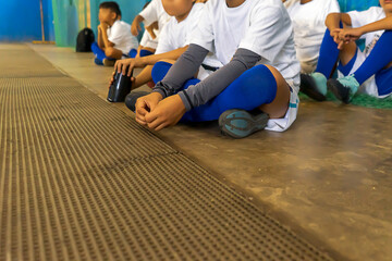 A team of unrecognizable latin soccer kids wait sitting on the floor in an indoor field.