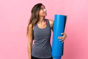 Young sport woman going to yoga classes while holding a mat isolated on pink background looking up while smiling