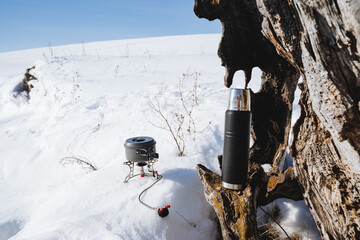 Thermos pan burner, camping camping utensils, old tree root, white snow, sunny weather.