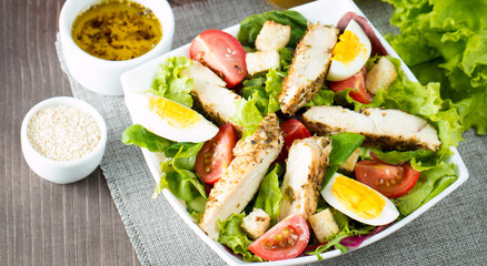 Fresh Caesar salad made of tomato, ruccola, chicken breast, eggs, arugula, crackers and spices. Organic ingredients in a white, transparent bowl on wooden background