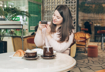 Pretty young girl sitting with a cup of coffee in a cafe. Lovely women enjoys coffee at a coffee shop. Girl blogger shooting food with cellphone.