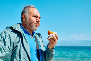 Bearded mature man at spring seaside eating hot palatable arancina (deep fried rice balls with...
