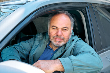 Happy smiling bearded mature man looking out the car window sitting behind the wheel of a prestigious car.