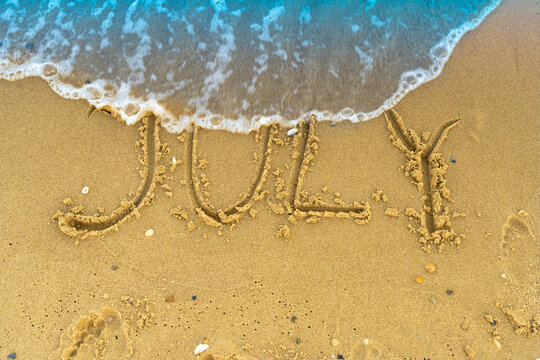 The word July is written on the sand of a beach by the sea. Washed off by a wave. The inscription disappears. End of summer concept. Back to school
