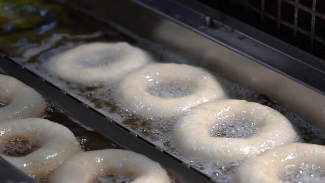 This video shows delicious hot and fresh donuts cooking in oil on factory conveyor belt on a busy day of food production.