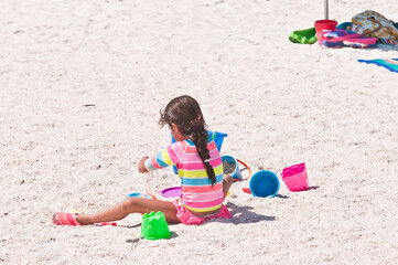 back view, medium distance, of a little girl in a colorful bathing suit, playing with colored, plastic toys on a sunny, sandy, tropical, beach