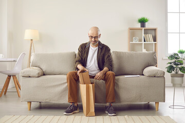 Customer opens brown paper bag he got from online store. Happy bald man sitting on couch in modern...