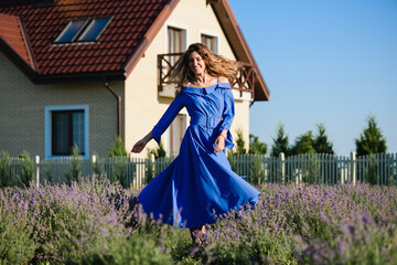 Fototapeta premium An attractive young positive woman in a blue dress is spinning, dancing in a lavender field. Country house in the background. Hair and dress are blowing in the wind.