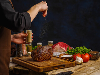 A professional chef prepares a ham on a wooden cutting board on a dark blue background. Vegetables, meat, spices on a wooden table. Recipes for meat dishes of restaurant and home cooking, restaurant.