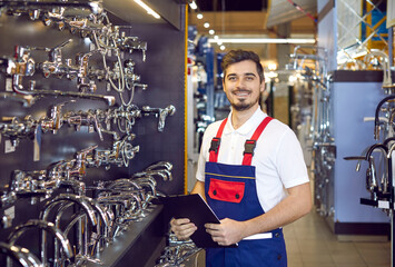 Portrait of a young man who sells faucets. Happy salesman in uniform standing in the aisle with modern water taps at a big hardware store or shopping mall - obrazy, fototapety, plakaty