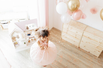 Little girl wearing pink dress having fun with golden pink confetti, balloons on background. Styled home birthday party
