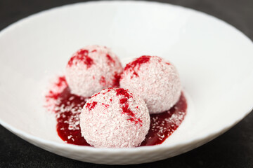 Homemade coconut balls, raw and healthy sugar free rafaello candies. Vegetarian energy sweets recipe. Round tasty dessert in bowl with strawberry sauce on dark background. Copy space, close up view.