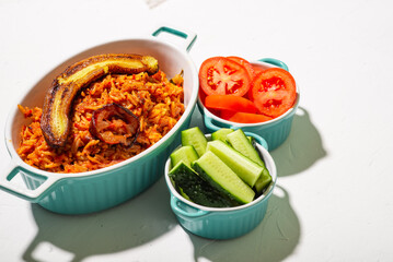 Jollof rice with fried banana. Fresh vegetables - tomato and cucumber. White background. Traditional Nigerian rice food with tomatoes, onions and spices.