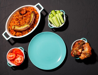 Jollof rice with fried banana, baked chicken. Rice with tomatoes, onions, spices. Traditional national Nigerian food. Dark background. View from above. Empty plate in turquoise color.