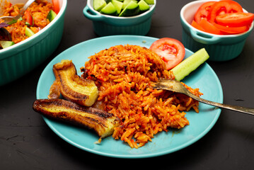 Jollof rice with fried banana on a plate. Rice with tomatoes, onions, spices. Traditional national Nigerian food. Dark background.