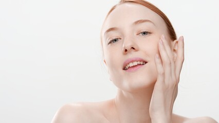 Young red-haired Caucasian woman with bare shoulders gently touches her cheek looking at camera on white background | Skin care comercial concept