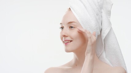 Young ginger Caucasian woman with a hair towel on her head gently touches cheek looking aside on white background | Smooth skin commercial concept