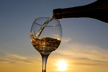 Pouring white wine from a bottle into the glass on sunset sky background. Celebration, romantic dinner with alcohol drink