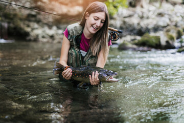 A young woman, who does sports fishing, fishes on a fast mountain river and holds a trout she caught in her hand.