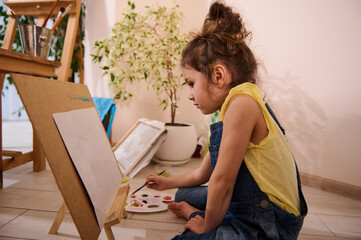 Adorable Caucasian child, cute little girl sitting on the floor at a wooden easel and drawing picture painting on canvas. Art class, creativity, children entertainment concept.