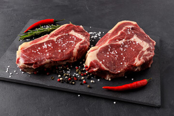 Two raw beef steaks on a cutting board with red hot peppers, rosemary and spices