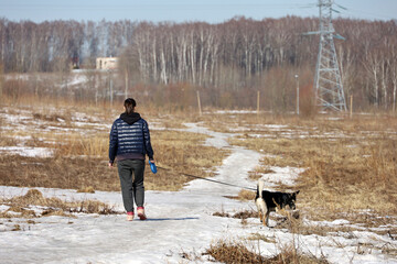 Girl in short coat walking a dog on a meadow. Suburban landscape in early spring season with dry grass and melting snow