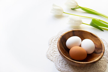 Brown and white raw chicken eggs in a wooden plate on a lace doily. Easter banner in sunshine with tulips on white background