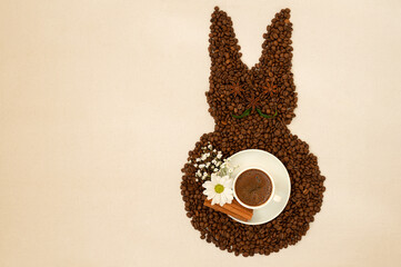 Creative top view flat lay of Easter bunny shape made of coffee roasted beans and cup of espresso on rustic table cloth. Rabbit is decorated with anise star, baby's breath flowers, daisy, cinnamon.