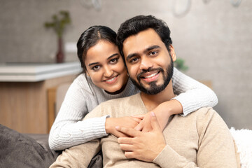 Portrait of lovely couple at home. Handsome man sitting on the comfortable couch, beautiful woman hugging his from the back. Spouses spend time together