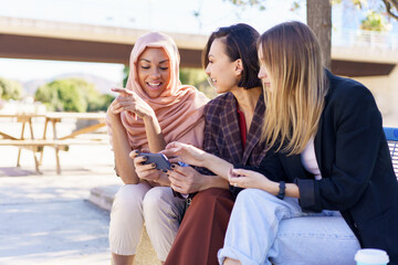 Positive multiethnic girlfriends sharing smartphone while resting on bench in park