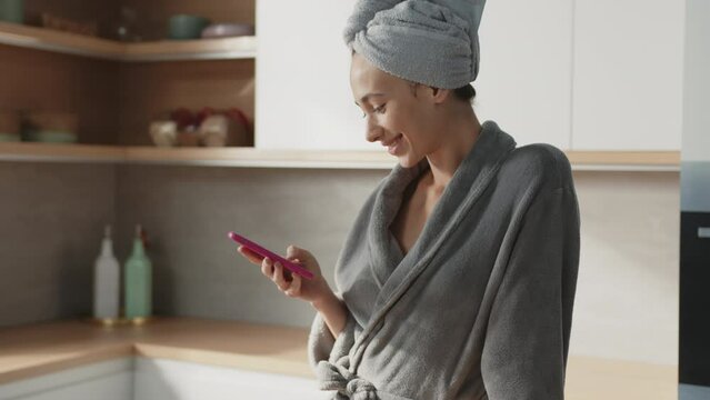 Attractive woman stand with towel on head use phone in the kitchen. Stand texing smiling, feel happy. Slow motion