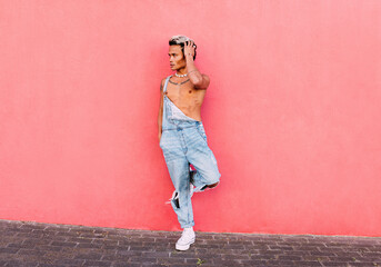 Stylish guy in casuals standing on one leg leaning on pink wall outdoors