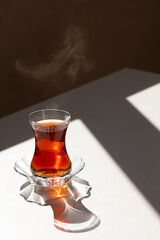 Close up of traditional Turkish tea cup on white table in sunlight with steaming vapors at black background. Relaxing tea break. Oriental way of pouring and serving tea in Turkey and Egypt.