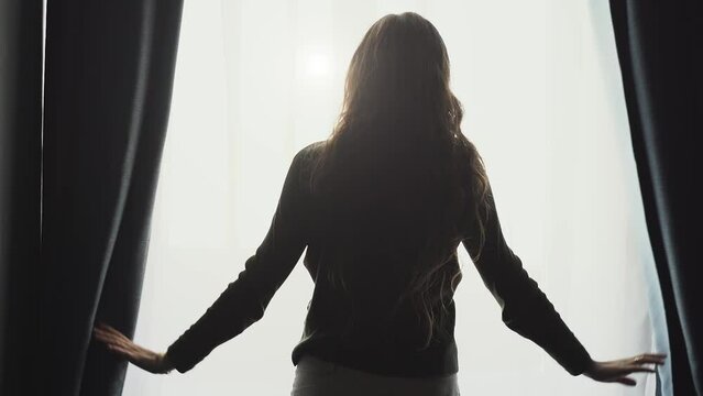 Young woman silhouette open curtains on window to morning light, rear view, slow motion.