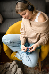 A young smiling girl is sitting in a yellow armchair and knitting a sweater with blue thread