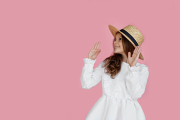 Front portrait of a cheerful little girl in cute dress and straw hat, looking at side, isolated on a pink background.