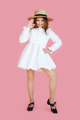 Full length image of a cheerful little girl in cute white dress, straw hat, looking at camera, on a pink background.
